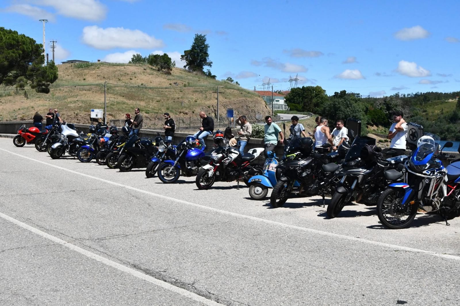 Fila de motas estacionadas ao longo de uma estrada junto a uma barragem, com motociclistas ao fundo
